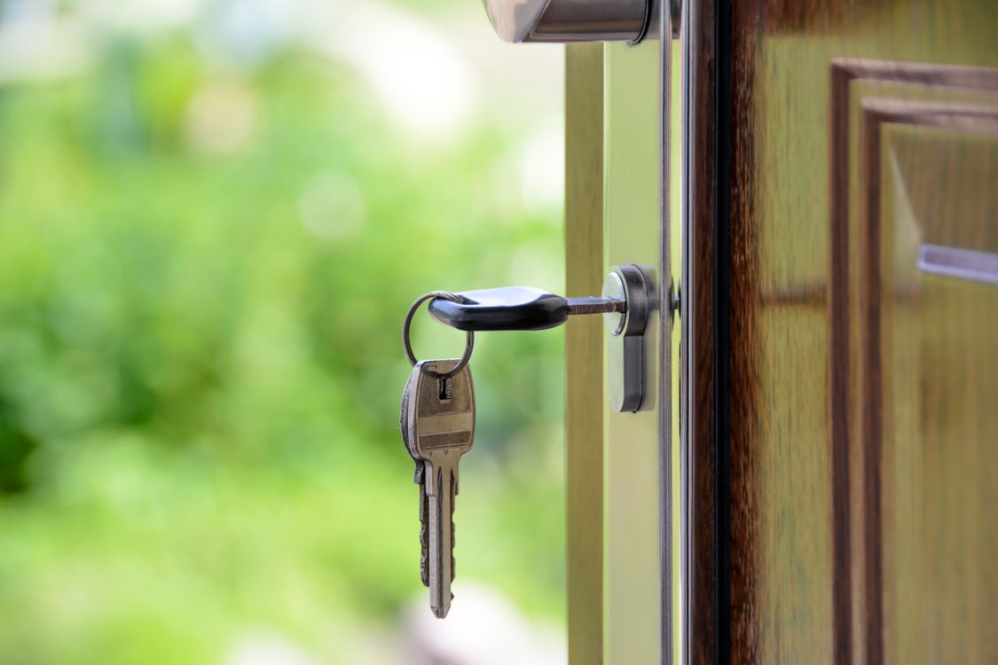 A close-up of a hand holding a single house key and one coin, with a modern home blurred in the background, illustrating the concept of low-fee real estate services.